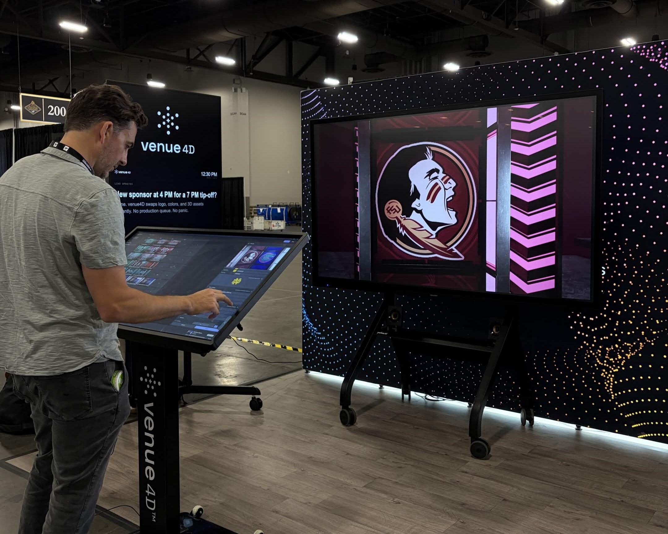 A man interacts with a large touch screen podium labeled "venue 4D" in an exhibition hall. The touch screen appears to control content on a large display monitor directly in front.