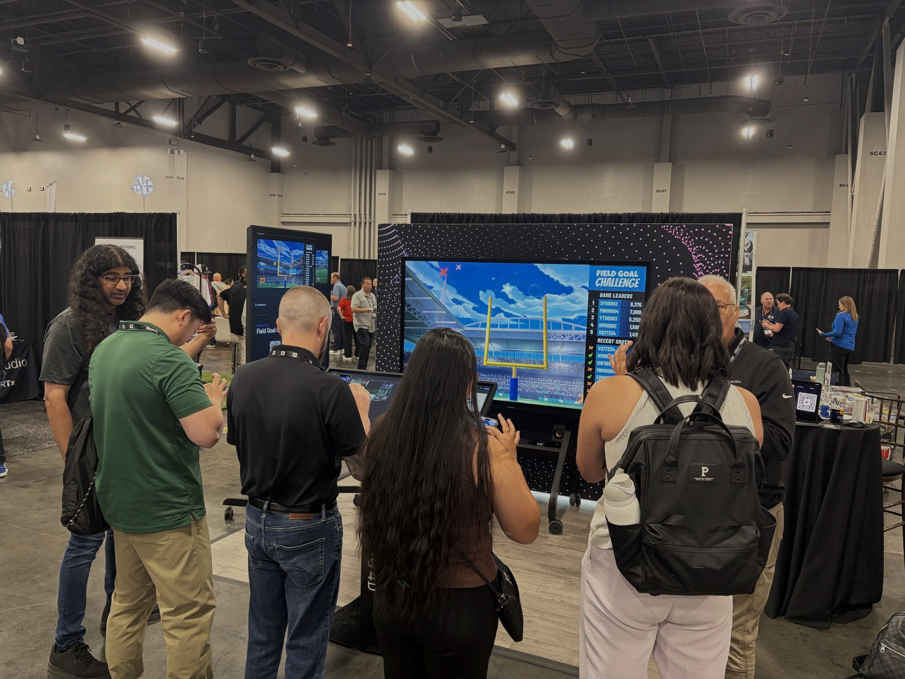 A group of people gathers around an interactive "Field Goal Challenge" game booth inside a spacious exhibition hall. Several individuals are focused on a control podium in the center, while a large display screen in the background shows an animated football stadium with a leaderboard on the right side.