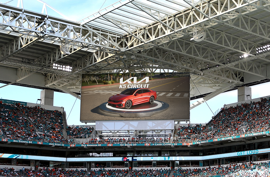 A stadium with a jumbotron displaying a racing game featuring a Kia car brand, with fans watching the interactive content.