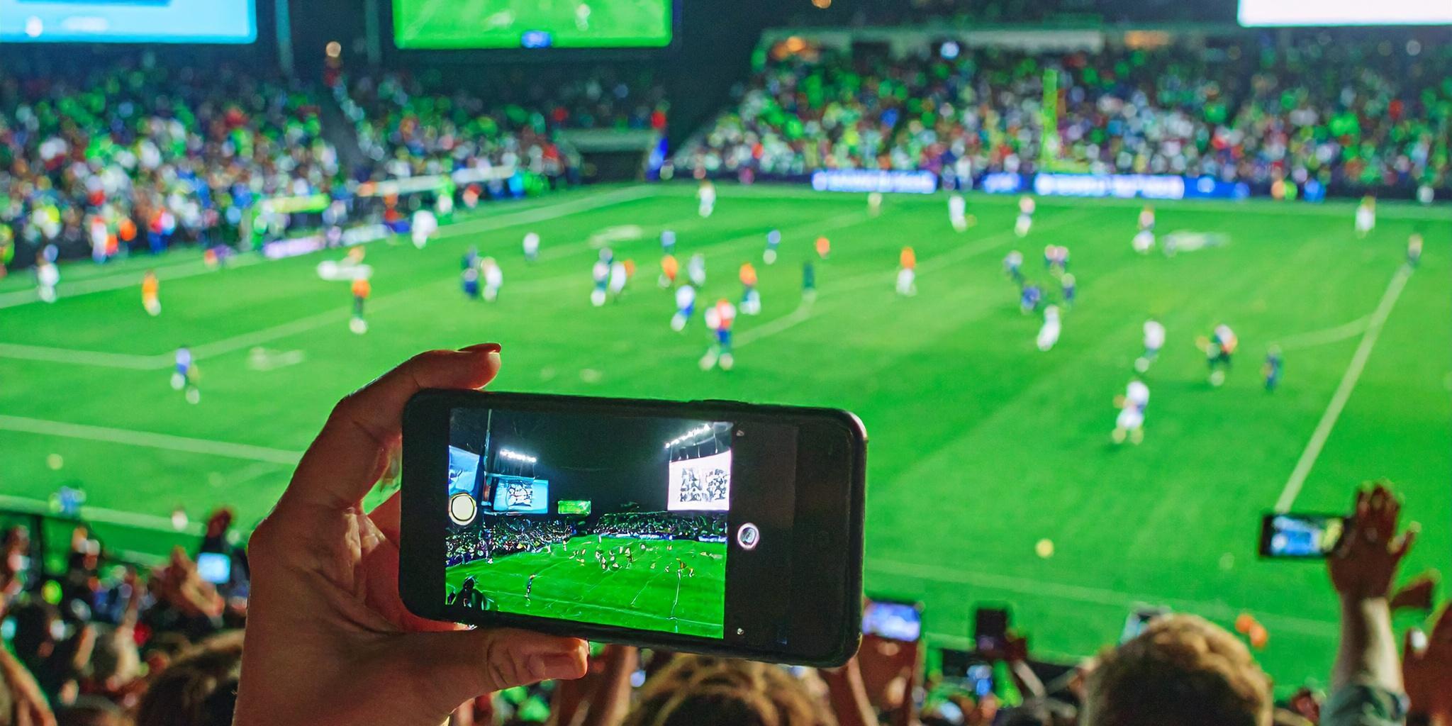 Fans capture soccer match on phones in a vibrant stadium with bright screens and live action.